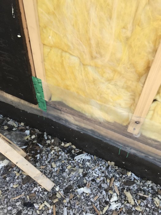 Bygningskonstruktion med gul isolering, plasticremse langs soklen og grus på jorden.