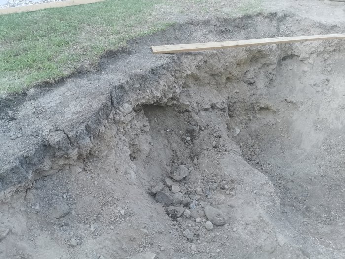 Grävd trädgårdsdamm på villatomt med ojämna kanter och synliga stenar och grus.