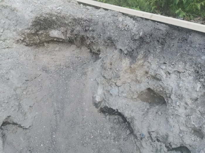 Nygrävd trädgårdsdammkant med synliga stenar och ojämn mark av sand och krossmaterial.