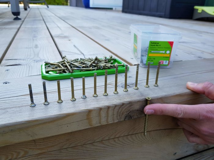 En hand som pekar på en avbruten skruv vid ett trädeck, med uppradade Essve Torpedo Max skruvar och en öppen förpackning i bakgrunden.