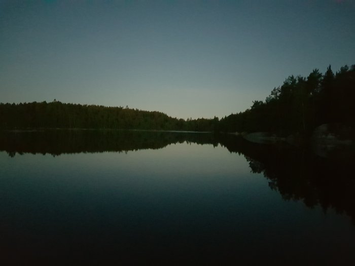 Sjö med stilla vatten vid skymningen, speglande skog och himmel på ytan.