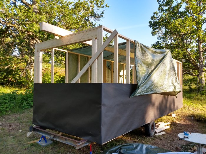 Påbörjat byggprojekt med uppresade väggstommar och nockbalk, täckt av presenning för skydd mot regn.