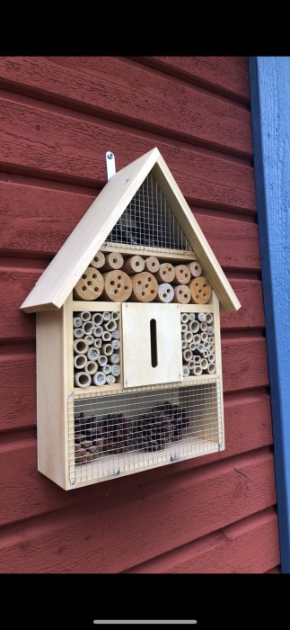 Insektshotell i trä med olika storlek rör hängande på en röd trävägg.