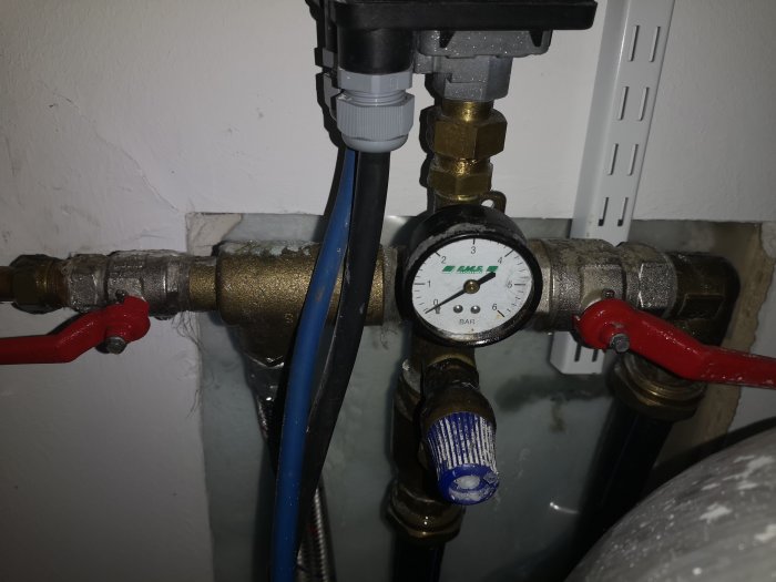 VVS-installation med manometer som visar 0, omgiven av rör och röda ventilhandtag.