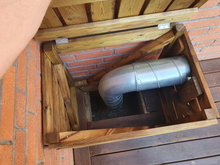 Förlängt ventilationssystemrör skyddat av en träbox med öppningsbar lucka och snehuv, monterat under en stängd altan.