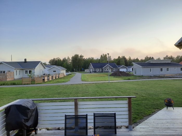 Nybyggt bostadsområde med gräsmatta, hus i bakgrunden, uppfart och en del av en träaltan i förgrunden.