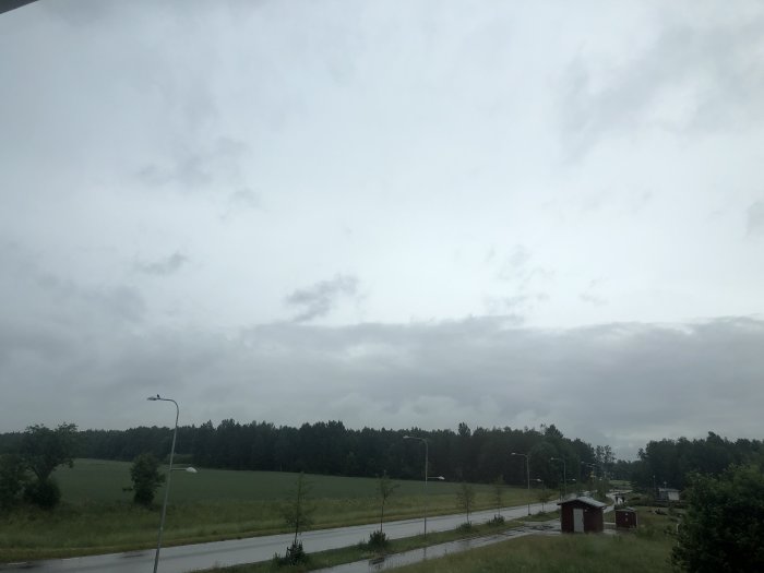 Molnig himmel över ett fält bredvid en väg med gatubelysning och små byggnader.