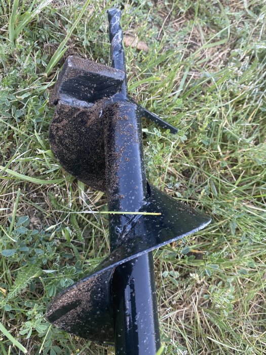 Spiralborr täckt med mörk jord ligger på gräsytan, indikerar byte av borr för att undvika lera.
