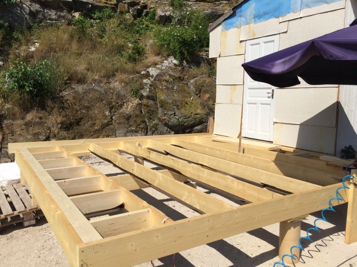 Timmerstomme under konstruktion med synliga reglar och bjälklag nära en byggnad och ett parasoll.