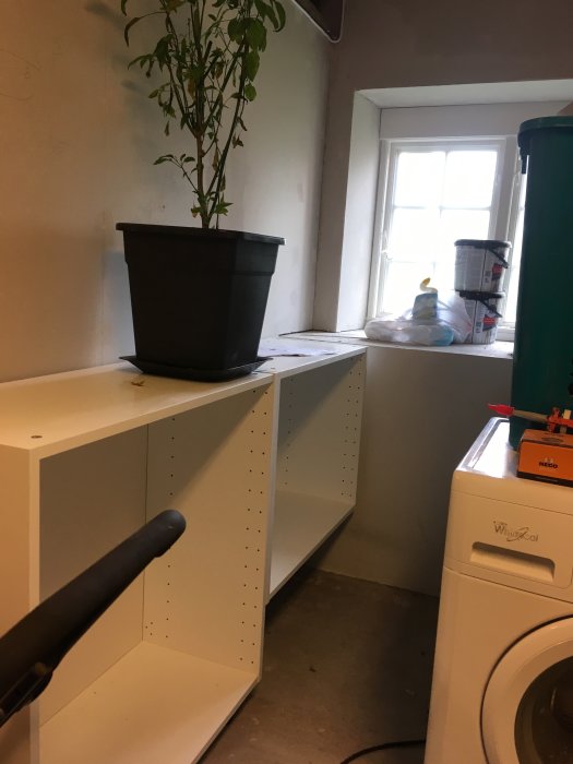 Två vita väggskåp av olika höjd utan dörrar, bredvid en tvättmaskin och en växt på skåpet.