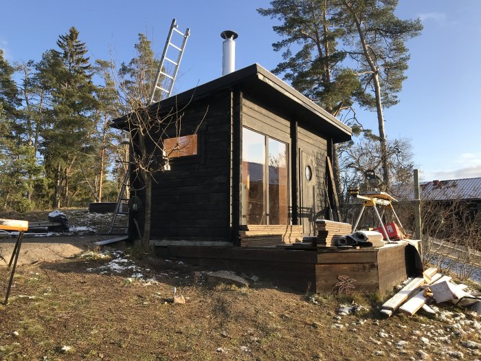 Nymålat svart utedhus med stora fönster, skorsten och byggmaterial synligt på en solig dag med lite snö.