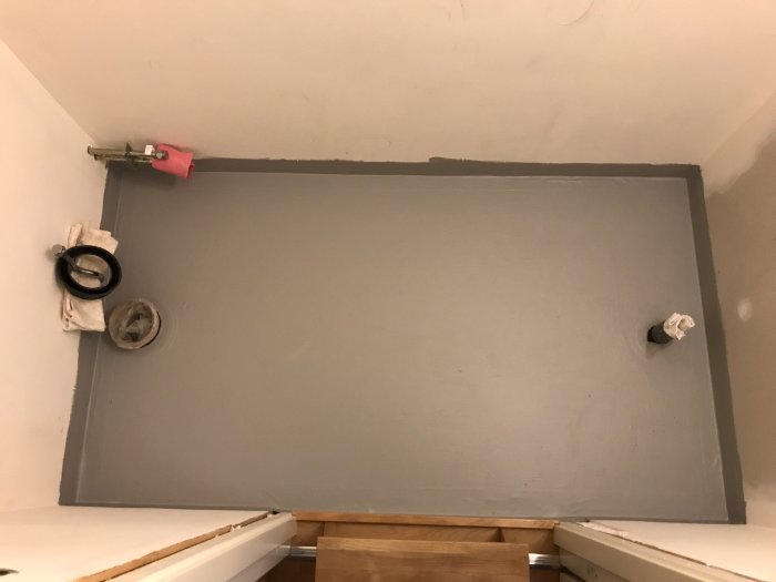 Renoverad gästtoalett med grå våtrumsmatta och manschetter runt avloppsrör, mönstrad remsa längs vägg-möte.