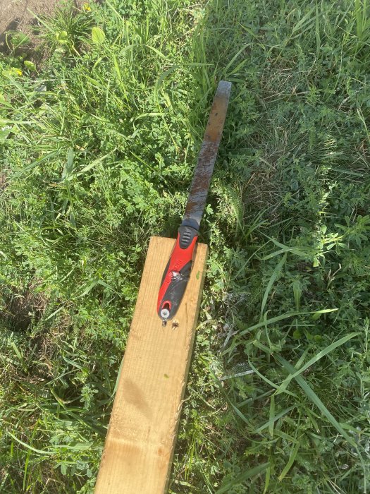 Sliten spett placerad på en träplanka med grön vegetation i bakgrunden, använd för grovarbete.