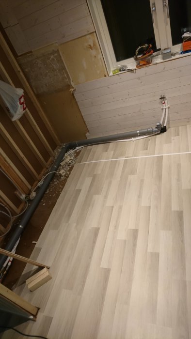 Renoveringsarbete med nytt golv och synligt avloppsrör längs väggen.