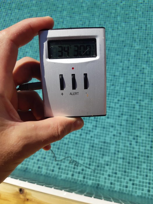 Hand håller en termometer som visar 34 grader Celsius framför en pool.