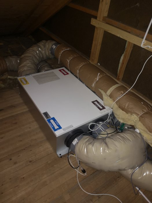 Ventilationsenhet på vinden med isolerade rör och elektriska kablar på trägolvet.