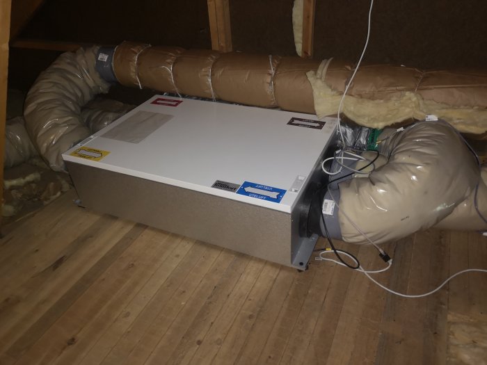 Ventilationsaggregat på vindsgolv med isolerade rör och elledningar i en mörk vindenmiljö.