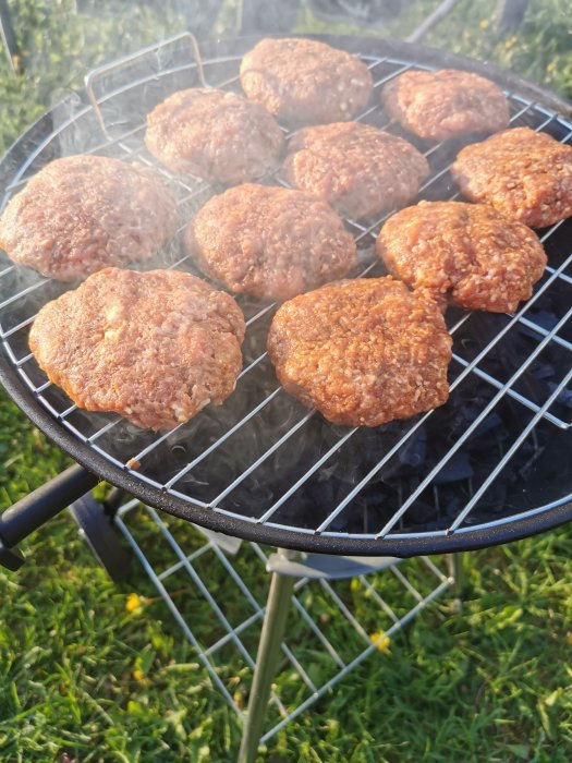 Hamburgare som grillas på en kolgrill utomhus med grönt gräs i bakgrunden.