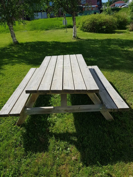 Träpicnicbord med bänkar på gräsmatta i solljus.