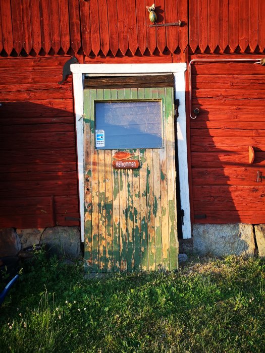 Gammal sliten träddörr med flagnande grön färg, halvt glasat övre fönster och "Välkommen"-skylt på en röd stugvägg.