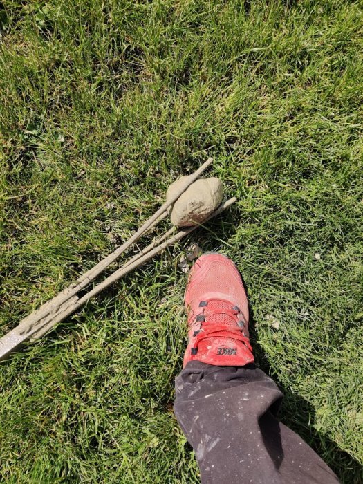 En persons smutsiga röd sneaker på gräset bredvid en hemmagjord stenfångare bestående av en träpinne och sten.