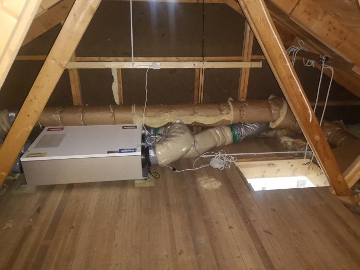 Ventilationsaggregat placerat på ett vindsbjälklag med synliga isolerade rör och en öppen vindslucka i förgrunden.