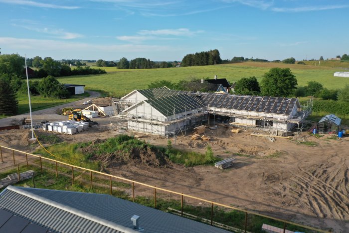 Hus under konstruktion med ställningar, byggmaterial och grävmaskin på landsbygd.