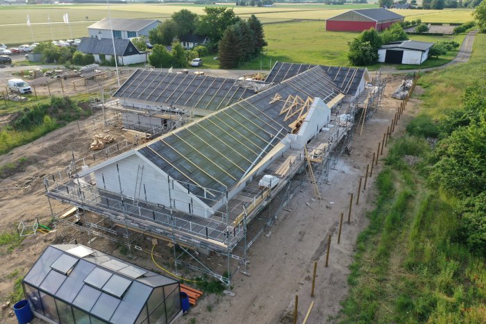 Flygbild över ett pågående husbygge med spikat takstolskonstruktion, säkerhetsställningar och omgivande landskap.