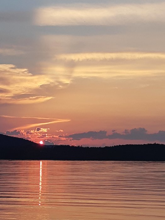 Solnedgång över vatten med solen som reflekterar på ytan och färgade moln i himlen.