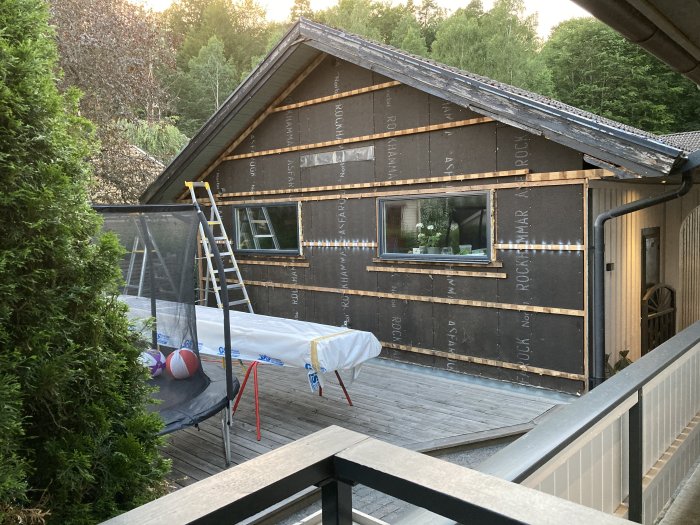 Hus under renovering med isolering exponerad, stege, täckt trädgårdsmöbel och studsande boll på terrass.