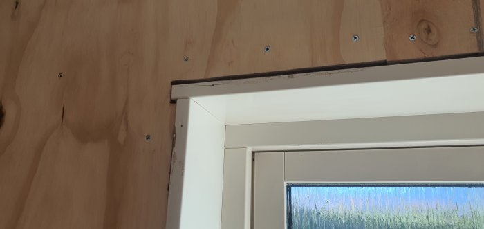 Plywoodplattor monterade ovanför ett vitmålat fönsterkarm, spår av tätningsskum synliga vid anslutning.