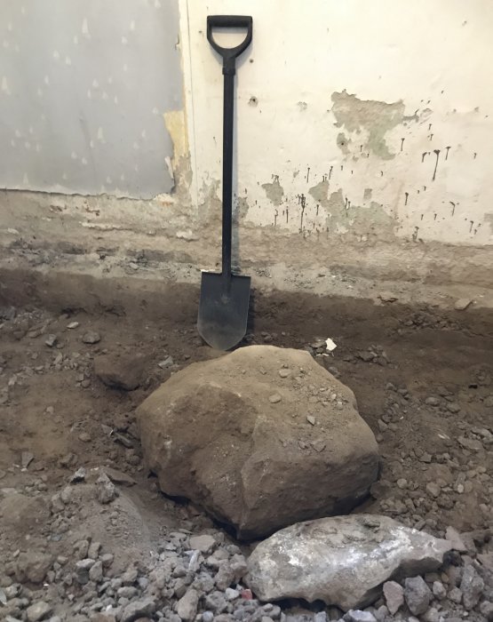 En spade står uppställd framför en stor sten bland grus och uppbrutet golv i en gillestuga under renovering.