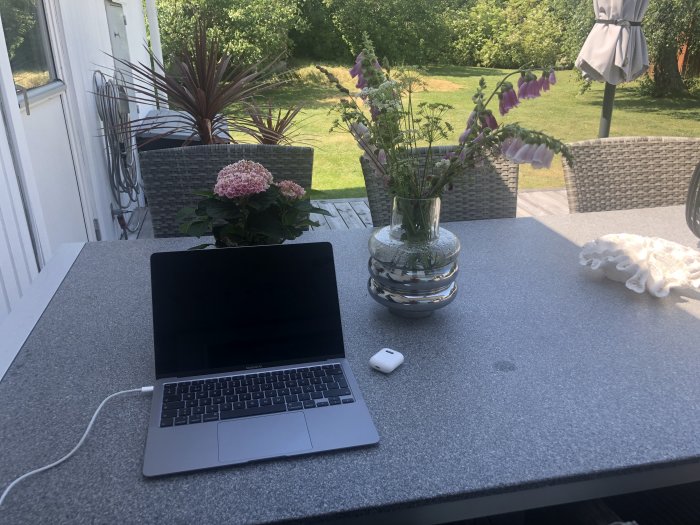 Arbetsplats utomhus med laptop på bord, blommor i vas, i en trädgård med grönska.