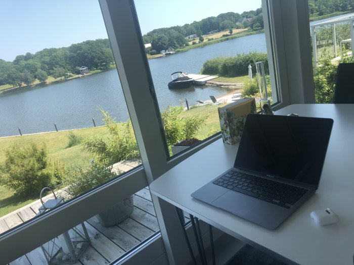 Arbetsplats med öppet fönster med utsikt över en sjö, grönska och en båt, i starkt solljus.