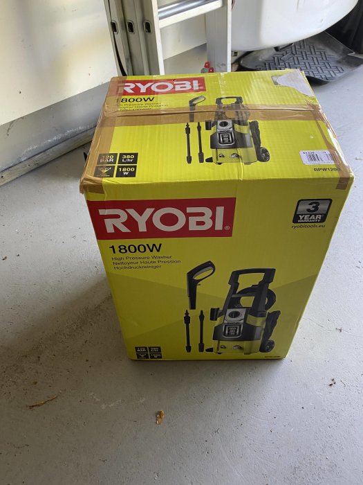 Förpackning av RYOBI högtryckstvätt 1800W i en kartong på ett garagegolv.