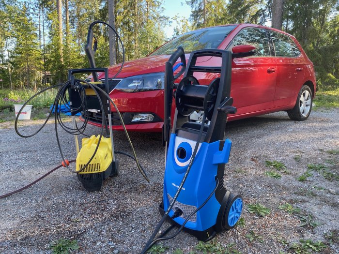 En ny högtryckstvätt ansluten till ett rött fordon på en grusuppfart med skog i bakgrunden, nära en äldre gul högtryckstvätt.