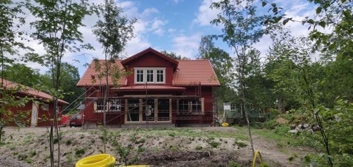 Nyrenoverat rött hus med plåttak och veranda i naturmiljö under blå himmel.