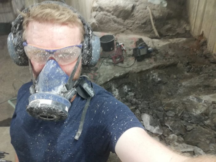 Person i skyddsutrustning tar selfie framför ränna i berg, med borrutrustning i bakgrunden.