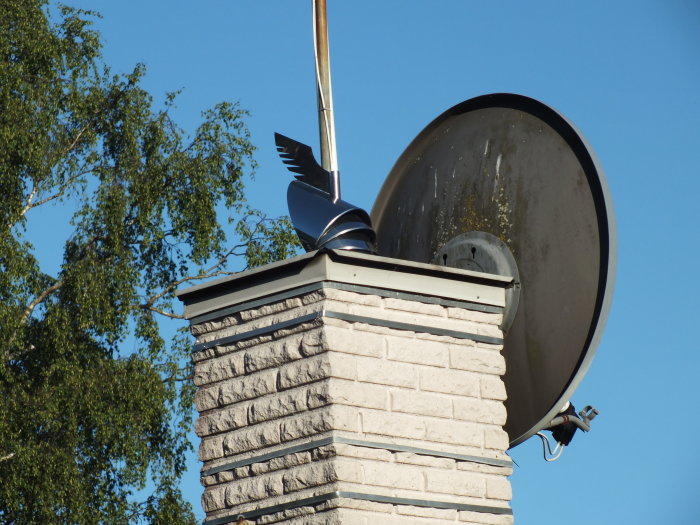 Parabolantenn med en plastfågel ovanpå på ett tegelstensskorsten mot en klarblå himmel.