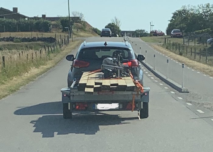 Bil som drar ett släp med träplattor och en motorcykel på en solig väg.