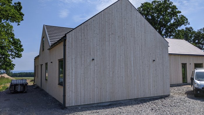 Vitaktig fasad på nybyggt hus med sluttande tak, väggarna lyser i solen, omgivet av natur.