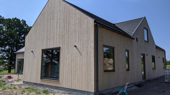Nybyggd husfasad med vertikal träpanel, stora fönster och svart tak, omgiven av en grusad gång och grön natur.