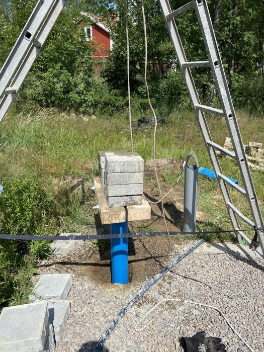 Byggplats med stenblock, blå rör i grävd hål, stegar och en byggnad i bakgrunden under solig himmel.