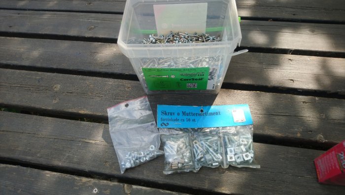 Olika skruv- och muttersortiment i förpackningar spridda på ett träbord, med fokus på en plastlåda med skruvar.