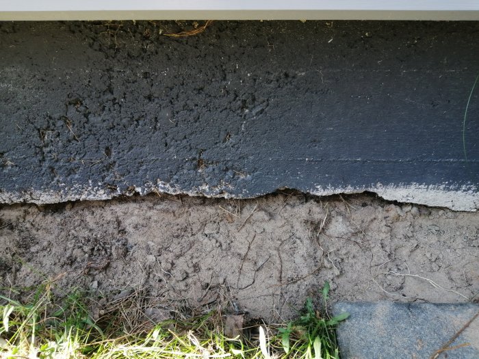 Skadad betongplatta vid basen av ett timmerhus, med synliga sprickor och tecken på erosion.