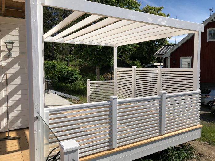 Nybyggd vit pergola med horisontella lameller, tjocka hörnstolpar och infällda överliggare i en trädgård.