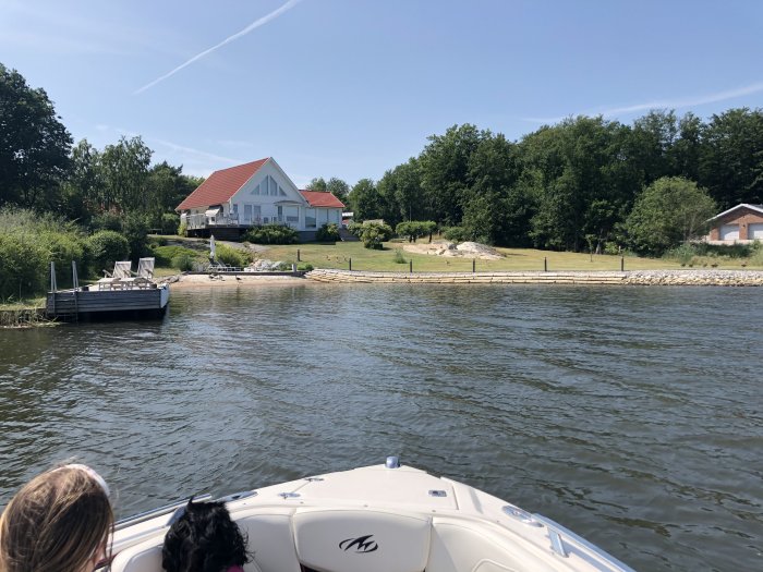 Vy från en båt mot ett hus med vita väggar och rött tak vid vattnet, brygga med sittbänkar framför huset.