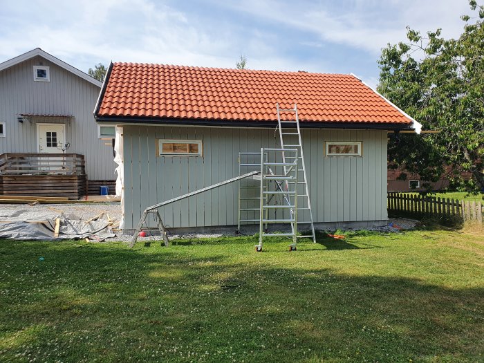 Nyinstallerat rött tegeltak på grått uthus med stege lutad mot taket, i en trädgård.