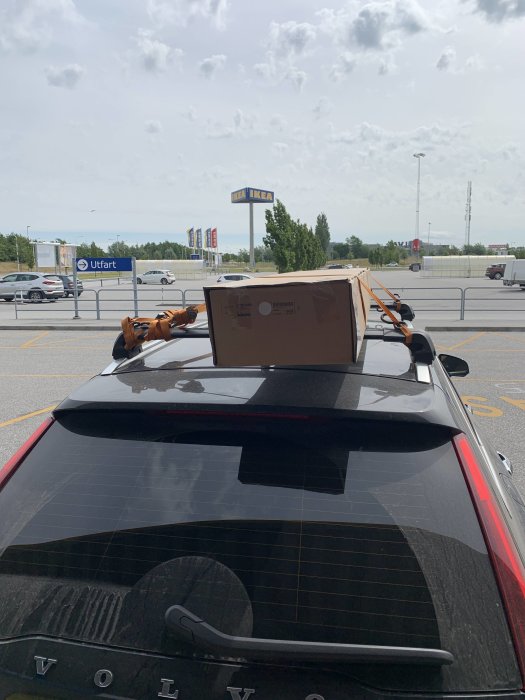 Bil med lastsäkrad kartong på taket framför IKEA, med spännband över kartongen och IKEA-skylt i bakgrunden.