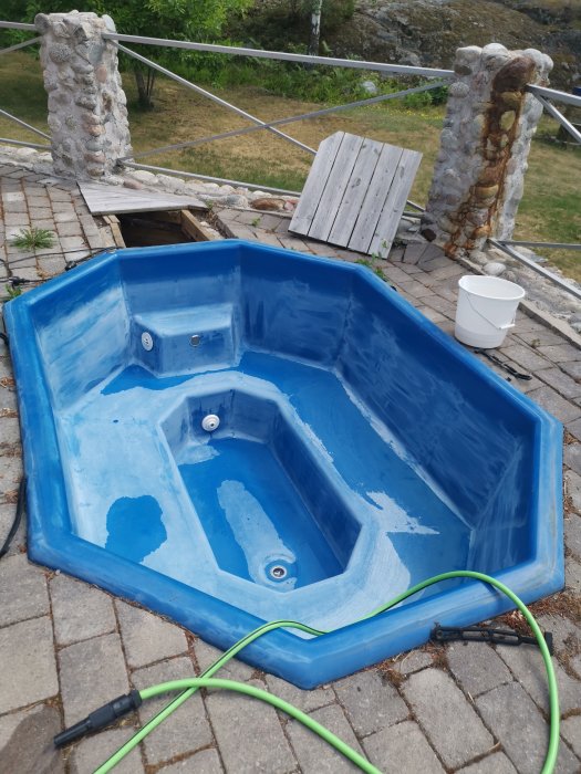 Tom inbyggd pool med blått skal på en stenlagd yta, sliten yta med slitage, med trädgårdsslang och hink vid sidan.